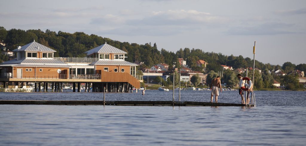 Bild på kallbadhuset i Ulricehamn med människor i förgrunden som badar från en brygga.