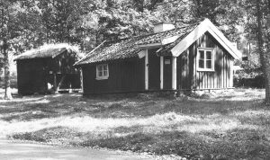 Svartvit bild på två gamla stugor. Den närmast i bild är låg och har vita knutar. 