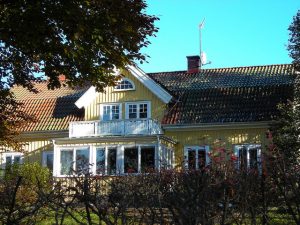 Bild på ett gult trähus med rött tegeltak och vita knutar. Huset har två våningar och en balkong.