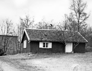 Svartvitt foto på en liten toparbyggnad med vita fönster och dörr. 