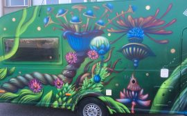 Bild på en husvagn målad i grön bakrund med konstnärliga figurer och växter.