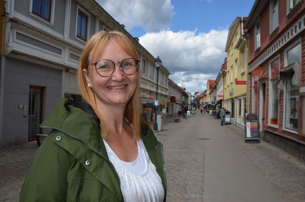 Bild på medelålders kvinna i grön jacka som leende står med gågatan i Ulricehamn i bakgrunden. Linda Larsson Järleklint på gågatan i Ulricehamn.