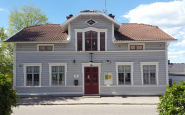 Bild på ungdomens hus, en grå träbyggnad i två våningar med röd dörr och vita fönster.