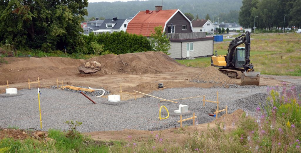 Bild på en tomt som håller på att markberedas av en grävskopa. ett färdigbyggt hus i bakgrunden.