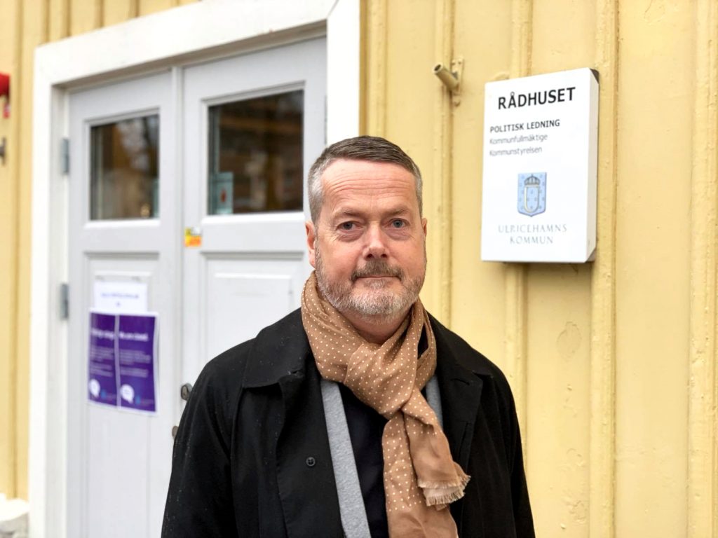 En bild på kommunchefen Gustaf Olsson, en man i 50-årsålderna som står framför en gul träbyggnad.