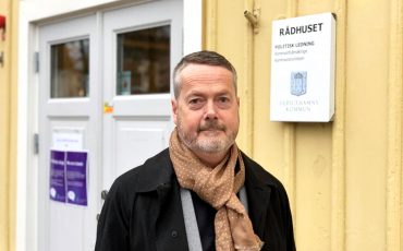 En bild på kommunchefen Gustaf Olsson, en man i 50-årsålderna som står framför en gul träbyggnad.