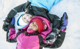 Två barn ligger i snön, ett har röd mössa och rosa overall, den andra har vit mössa och svartblå overall. Båda ler.