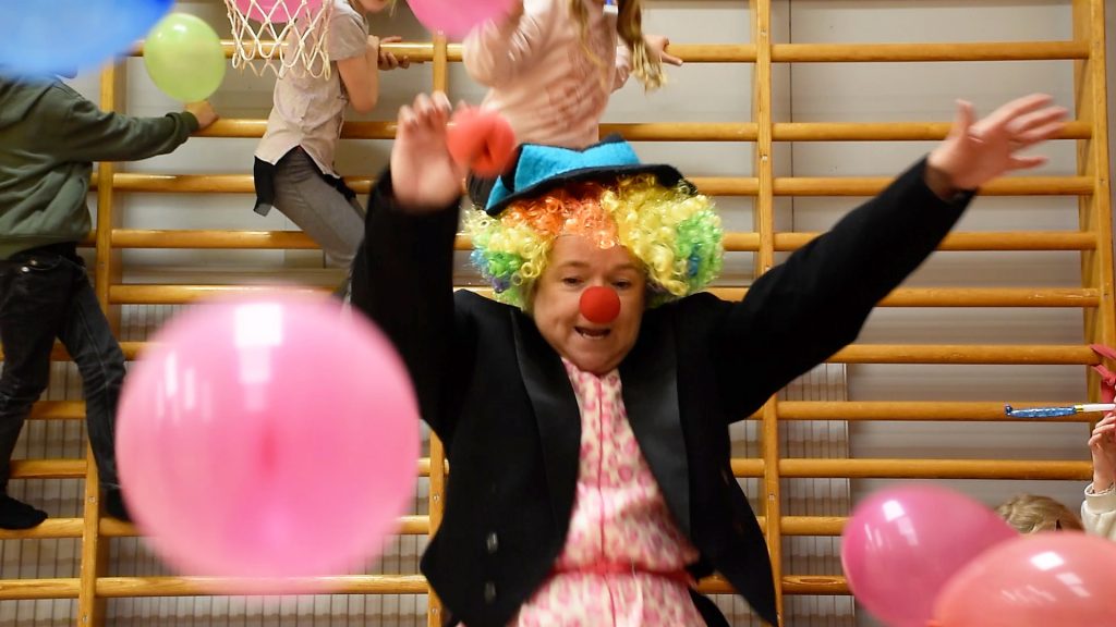 En clown i regnbågsfärgad peruk åker rutschkana nerför en bänk lutad mot en ribbstol i en gympasal. I luften flyger ballonger och i bakgrunden klättrar barn på ribbstolarna.