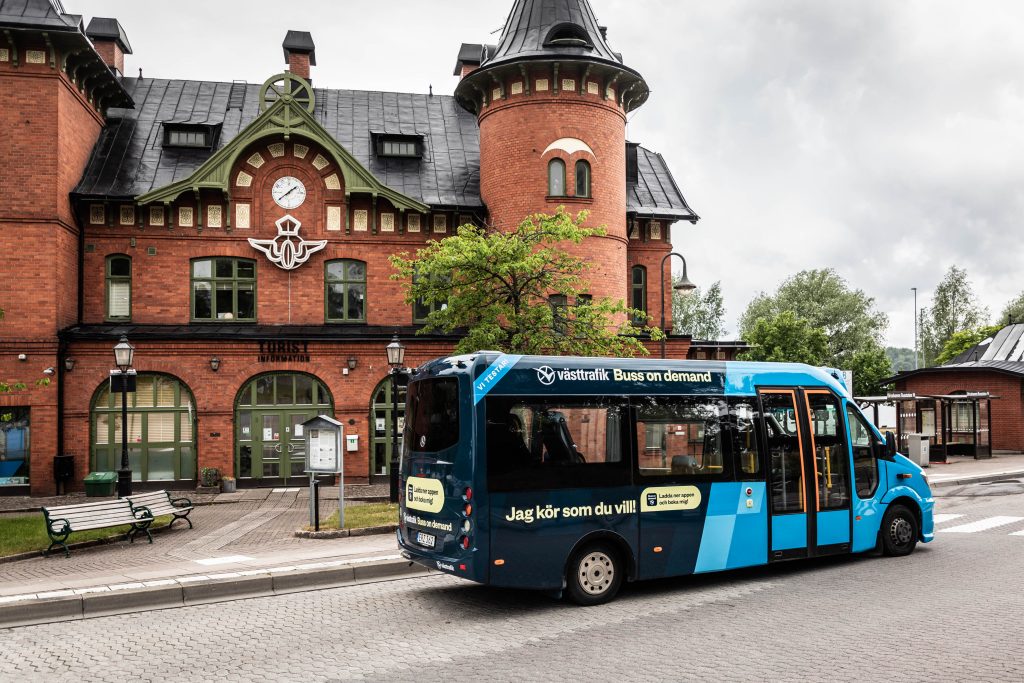 En blåsvart buss åker på en busshållplats. I bakgrunden finns ett stationshus i rött tegel.