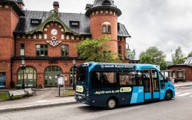 En blåsvart buss åker på en busshållplats. I bakgrunden finns ett stationshus i rött tegel.