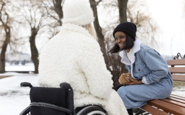 En kvinna sitter på en bänk och en annan kvinna sitter i en rullstol. Kvinnan i rullstol sitter med ryggen mot fotografen. Kvinnan på bänken lutar sig fram och skrattar