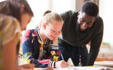 En lärare hjälper två elever i ett klassrum. Eleverna sitter och skriver och läraren står bakom och kollar på deras arbete.