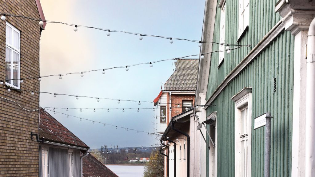 Linspänd belysning mellan brandgult tegelhus och grönt trähus med vita knutar.