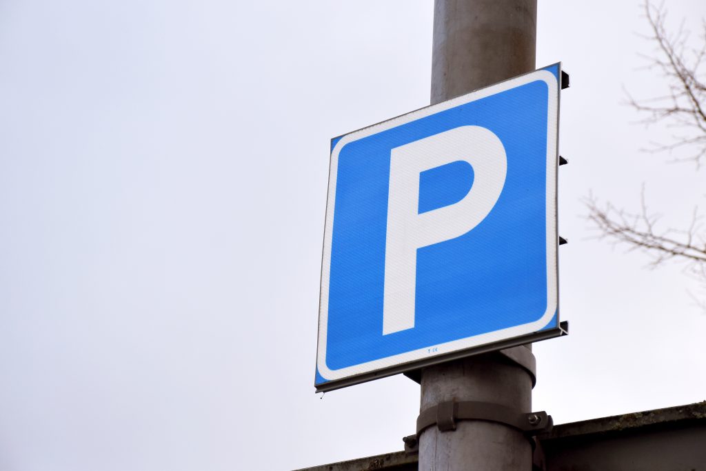 En närbild på en blå skylt med ett stort P på. Skylten sitter på en stolpe vid en parkering.