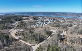 En väg som går genom ett område med skog. Bredvid ligger ett bostadsområde med villor, i bakgrunden syns sjön Åsunden.